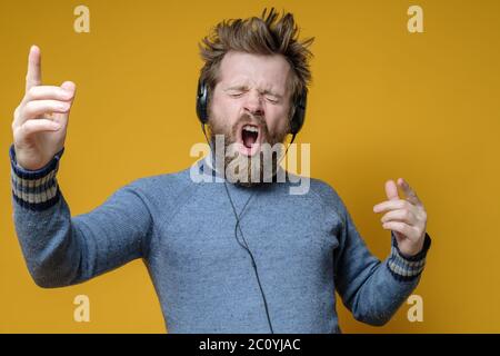 Un hipster satisfait dans un ancien pull écoute la musique sur les écouteurs et chante fort, émotionnellement le long, faisant des gestes de main. Banque D'Images