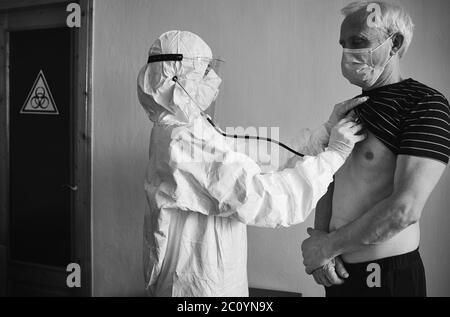 Médecin en costume de protection personnelle examinant le patient âgé de sexe masculin à la maison à l'aide d'un phonendoscope, en vérifiant la poitrine, debout au mur, panneau de danger biologique sur la porte, noir et blanc, concept COVID-19 Banque D'Images