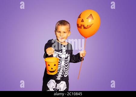 Mignon petit garçon dans le costume d'Halloween de squelette et orange ballon tenir seau de bonbons sur fond violet Banque D'Images