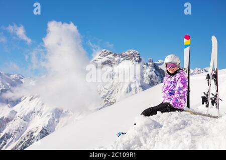 Bonne fille dans la tenue de ski, casque et masque assis dans la neige sur le sommet de montagne regarder la caméra Banque D'Images