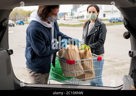 Vue sur le coffre d'un jeune couple dans des masques chargement des sacs dans le coffre après le shopping dans un supermarché Banque D'Images