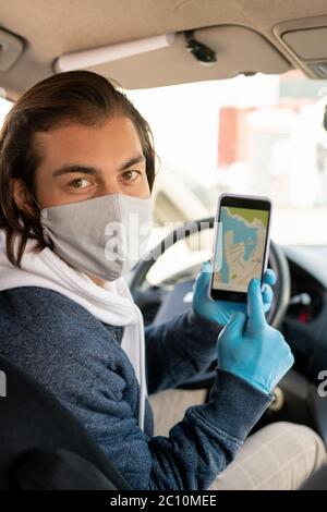 Portrait du chauffeur de taxi brunette dans un masque et des gants pointant sur la carte en ligne sur l'écran du smartphone tout en demandant au passager de l'endroit de destination Banque D'Images