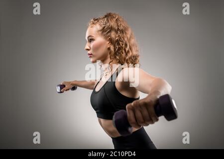 Vue latérale du jeune sportif en combinaison noire de levage des haltères et de respiration pendant l'exercice physique sur fond gris Banque D'Images