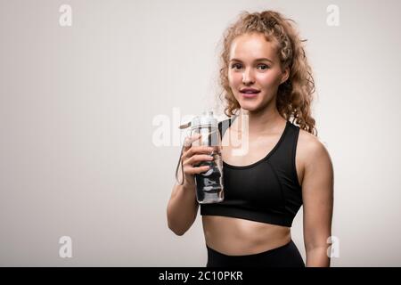 Bonne jeune sportswoman en casque d'activité noir va boire de l'eau de bouteille en plastique après un entraînement difficile en se tenant devant l'appareil photo Banque D'Images