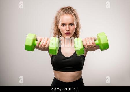 Jeune sportif actif en combinaison noire debout devant l'appareil photo et regardant vous pendant que vous faites de l'exercice avec des haltères vertes Banque D'Images