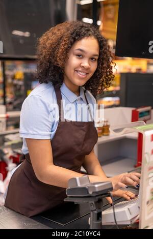 Joyeux jeune caissière de course mixte avec des cheveux ondulés foncés vous regardant en vous tenant debout près du lieu de travail et en appuyant sur le bouton de la cashbox Banque D'Images