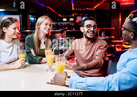 Deux joyeux gars multiethniques discutant des moments curieux de la dernière partie de bowling tandis que leurs amies avec des boissons assis à proximité Banque D'Images
