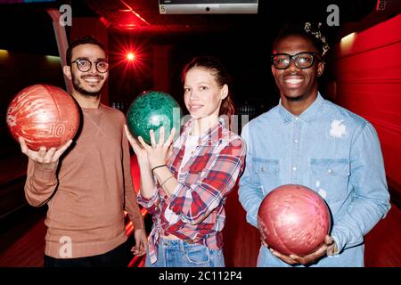Jolie jeune femme et deux types interculturels avec des sourires crasseux tenant des boules de bowling tout en jouant ensemble dans un club de divertissement Banque D'Images