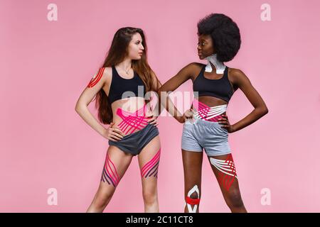 Enregistrement de kinésiologie. Deux jeunes athlètes féminines sur fond rose avec bande kinésiologique sur le cou, l'épaule, le khee, le ventre et les hanches. Perte de graisse, cellulite Banque D'Images