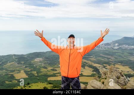 Homme en veste orange debout au sommet des montagnes avec la vue sur la mer et criant avec bonheur Banque D'Images