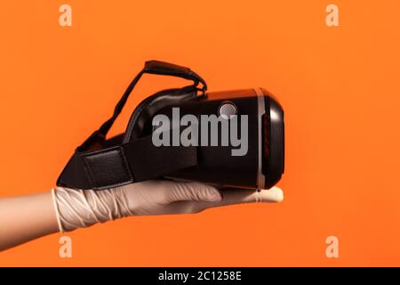 Vue latérale du profil gros plan de la main humaine avec gants chirurgicaux blancs tenant un casque de réalité virtuelle. Prise de vue en studio, isolée sur fond orange. Banque D'Images