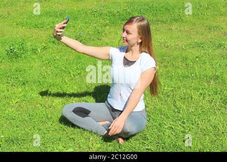 Une belle jeune fille blanche dans un t-shirt blanc et avec de longs cheveux assis sur l'herbe verte, sur la pelouse et prend un selfie. Banque D'Images