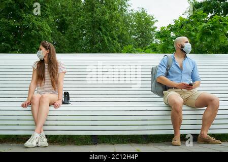 Homme et femme regardant dans différentes directions assis sur les extrémités opposées d'un banc en gardant la distance l'une de l'autre pour éviter la propagation du coronavirus Banque D'Images