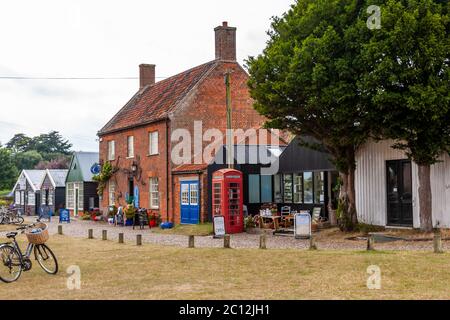 Sur un square de village dans l’East Suffolk se dressent un cottage en brique au toit de tuiles rouges, un café vêtu de noir avec de grandes fenêtres et une cabine téléphonique rouge sous un arbre étalant ses branches ; plusieurs vélos s’appuient au bord de la route. À gauche, une rangée d’étals peints en sombre bordent le square. La scène se situe dans la paroisse de Henstead with Hulver Street, East Suffolk. Banque D'Images