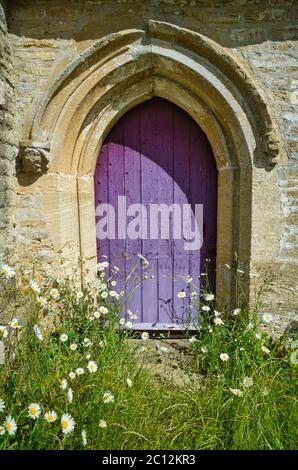 Ancienne porte en bois violet située dans l'église Cotswold avec des pâquerettes qui poussent en premier plan Banque D'Images