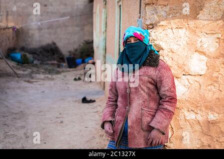 Portrait d'une femme berbère nomade avec son visage couvert, penchée sur un mur de briques de boue dans un simple village berbère dans la province de midelt, au maroc Banque D'Images