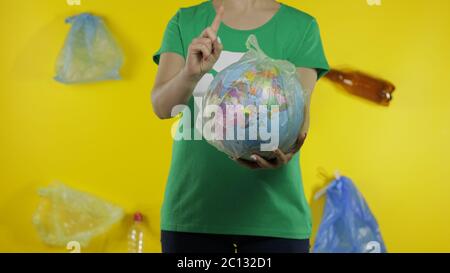 Femme imreconnaissable militante fille dans t-shirt avec logo de recyclage avec globe terrestre dans cellophane paquet. Fond jaune avec sacs, bouteilles. Sauver l'environnement écologique. Pollution par nature des déchets plastiques Banque D'Images