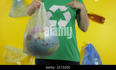 Femme imreconnaissable militante fille dans t-shirt avec logo de recyclage avec globe terrestre dans cellophane paquet. Fond jaune avec sacs, bouteilles. Sauver l'environnement écologique. Pollution par nature des déchets plastiques Banque D'Images