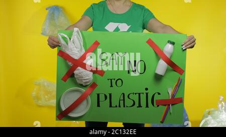 Femme imreconnaissable militante dans un t-shirt avec logo de recyclage tenant le message de protestation affiche dire non à plastique. Fond avec sacs de cellophane, flacons. Pollution par les déchets plastiques de l'environnement Banque D'Images