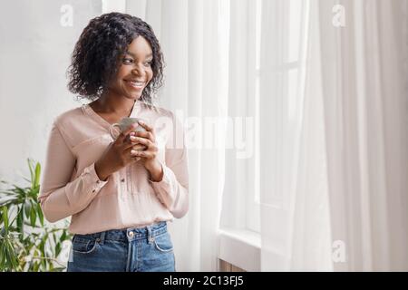 Parfait pour commencer la journée. Petite fille afro-américaine pensive buvant un café du matin contre la fenêtre et rêvant de jour, panorama avec espace libre, vue latérale Banque D'Images
