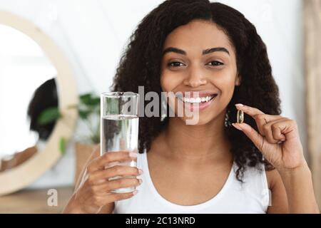 Vitamines et compléments alimentaires. Femme noire souriante tenant une capsule Omega-3 et de l'eau Banque D'Images