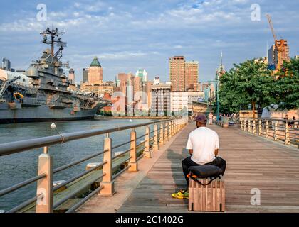 Un homme non identifié est assis sur un banc au quai Pier 84, dans la soirée. L'horizon de Midtown Manhattan et l'USS Intrepidaare en arrière-plan. Banque D'Images