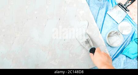 Spatule en métal à la main met du mastic sur le plan de travail, un pot de ciment, un pinceau pour la peinture molaire, un chiffon bleu et un revêtement de sol en cellophane bleu. Auto-réparation et décoration Banque D'Images