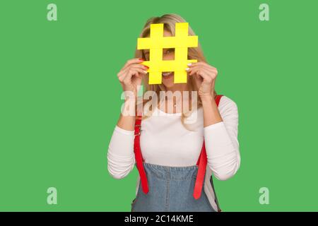 Commentaires de blog, tendances Internet. Portrait de belle femme positive dans des combinaisons en denim stylées regardant à travers le symbole hashtag, la promotion de la matière virale i Banque D'Images