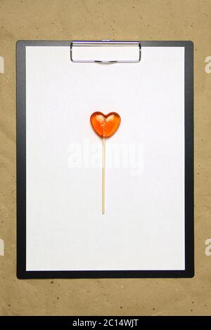 La tablette avec un clip pour le clip de papier avec une feuille blanche a4 repose sur l'arrière-plan du papier brun froissé. Sur le dessus se trouve un lollipop de caramel de rouge orange en forme de coeur. Place pour le texte et la mise en page pour la conception. Banque D'Images