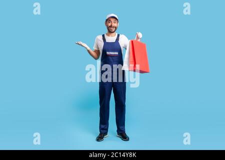 Portrait complet de courrier surprise en uniforme tenant des sacs d'achats et l'air choqué, livrant colis avec des marchandises commandées en ligne à la mode Banque D'Images