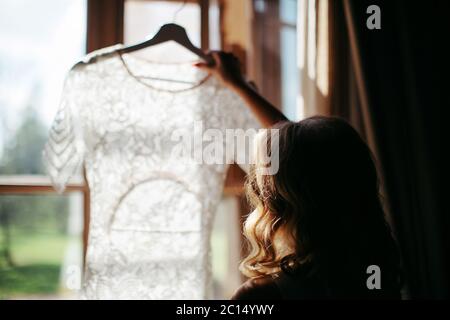 Femme blonde avec cheveux bouclés tenue et regardant la belle robe de mariage blanche sur un cintre, debout près de la fenêtre dans sa maison, vue arrière, daylig Banque D'Images