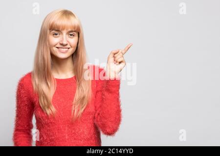 Bonne jeune blonde gaie femme dans un chandail rouge surprise montrant le produit, montrant à vide espace de copie avec son doigt, isolé sur fond gris. Banque D'Images