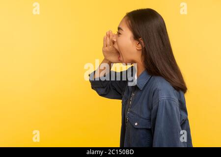 Attention, tout le monde écoute! Vue latérale de la fille en colère dans une chemise en denim criant dans les mains en forme de mégaphone, annonçant message négatif, voix forte. i Banque D'Images