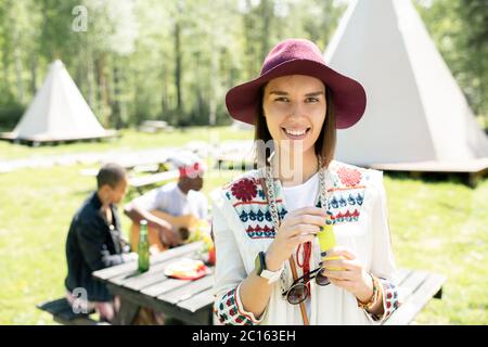 Portrait de la bonne fille hippie attrayante dans beau chapeau debout contre la table des amis au camping du festival Banque D'Images