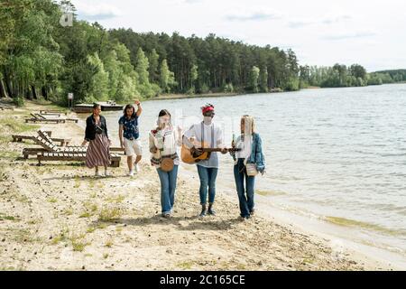 Groupe de jeunes amis multiethniques positifs qui s'amusent à se promener sur la plage tandis que le gars noir joue de la guitare en mouvement Banque D'Images