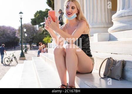 Belle fille blonde souriante avec masque facial après avoir rouvert le verrouillage - elle apprécie sa marche en s'amusant, en train de chatter avec des amis, en prenant selfies outsi Banque D'Images