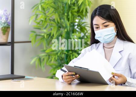 Portrait d'une femme asiatique médecin porter un masque de protection s'asseoir dans sa salle de bureau dans la clinique de l'hôpital et lire sur le dossier de patience avant de vérifier patie Banque D'Images