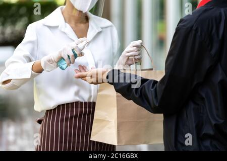 Serveuse vaporisant l'alocohol sur la main de l'homme de délivrance avant de donner le sac d'épicerie de boulangerie pour le nettoyage et l'hygiène comme nouvelle norme pour le service de livraison de nourriture W Banque D'Images