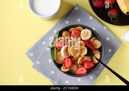 Petites crêpes tendance pour le petit déjeuner avec fraise et banane dans un bol sombre sur fond jaune et une tasse de lait, vue du dessus Banque D'Images
