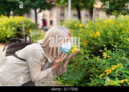 Femme essayant de sentir une fleur tout en portant un masque dans le parc, concept de pandémie de coronavirus Banque D'Images