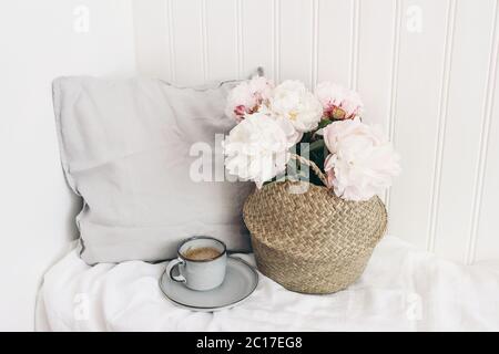 Scène féminine de mariage ou d'anniversaire. Bouquet de pivoines roses dans un panier de paille, une tasse de café, un oreiller en lin. Fond de mur en bois blanc Banque D'Images