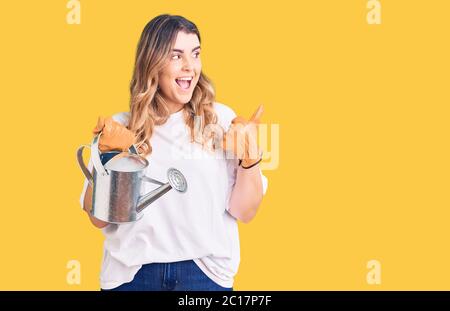 Jeune femme caucasienne portant des gants tenant l'arrosage peut pointer le pouce vers le côté souriant heureux avec la bouche ouverte Banque D'Images