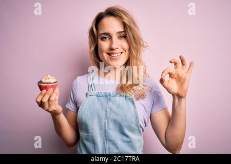 Jeune belle femme blonde eatimg chocolat cupcake sur fond rose isolé faire signe ok avec les doigts, excellent symbole Banque D'Images