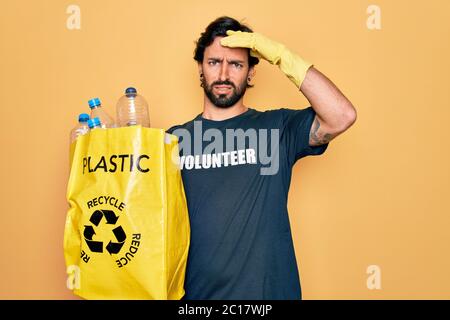 Beau volontaire hispanique portant un t-shirt de bénévolat sac de maintien avec des bouteilles en plastique stressé avec la main sur la tête, choqué par la honte et la surrp Banque D'Images