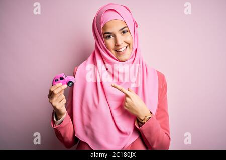 Jeune fille portant le hijab musulman tenant le jouet de petite voiture sur un fond rose isolé très heureux pointant avec la main et le doigt Banque D'Images