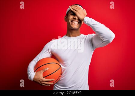 Jeune sportif afro-américain élégant tenant le ballon de basket-ball sur fond rouge stressé de main sur la tête, choqué par la honte et le visage surprise Banque D'Images