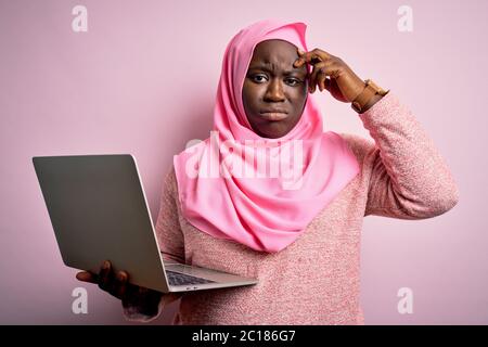 Femme afro-américaine de taille plus portant le hijab musulman utilisant un ordinateur portable sur fond rose inquiet et stressé au sujet d'un problème de main sur le front, ne Banque D'Images