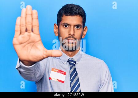 Jeune homme latin portant l'autocollant avec bonjour mon nom est message avec main ouverte faisant signe d'arrêt avec une expression sérieuse et confiante, geste de défense Banque D'Images