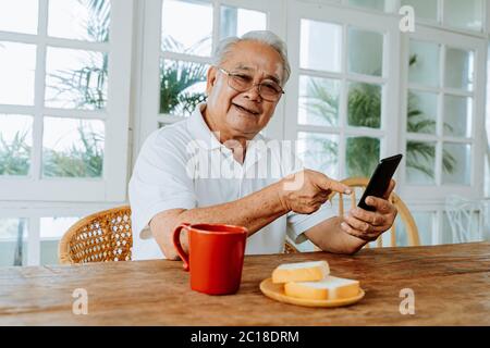 Joyeux homme asiatique âgé souriant pour l'appareil photo et pointant vers le téléphone portable tout en étant assis à table avec une boisson chaude et du pain le matin à la maison Banque D'Images