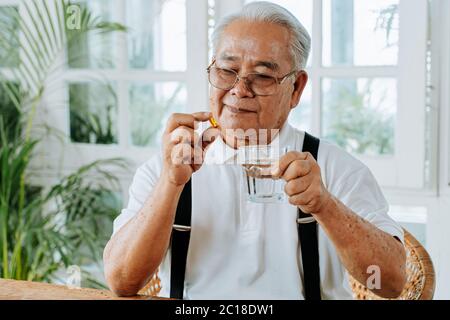 Homme asiatique senior avec une tasse d'eau et de pilule prenant remède dans la chambre confortable à la maison. Vieux mâle prenant des médicaments Banque D'Images